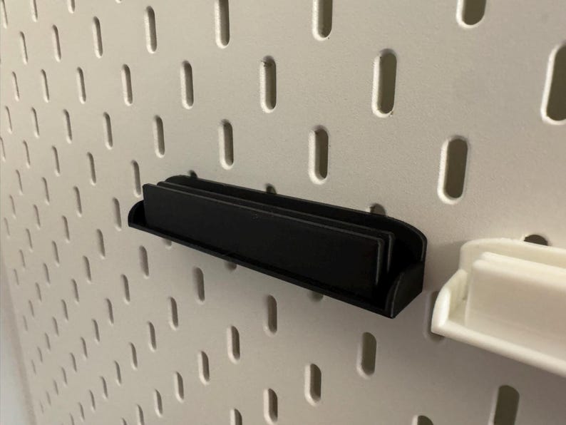 Pode incluir: Uma prateleira preta para painel perfurado &eacute; montada em um painel perfurado branco. A prateleira &eacute; retangular com uma frente curva e foi projetada para guardar itens pequenos. O painel perfurado tem orif&iacute;cios ovais espa&ccedil;ados.