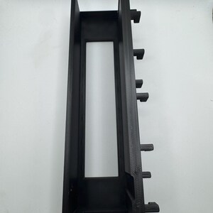 Peut inclure: Composant en plastique noir de forme rectangulaire, avec une d&eacute;coupe centrale et plusieurs languettes saillantes sur un c&ocirc;t&eacute;. L'objet a une finition mate et semble imprim&eacute; en 3D. Il s'agit probablement d'un &eacute;l&eacute;ment structurel ou de montage.