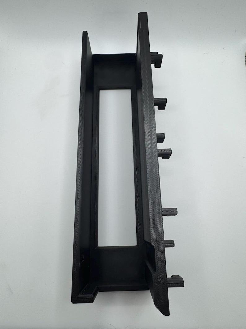 Peut inclure: Composant en plastique noir de forme rectangulaire, avec une d&eacute;coupe centrale et plusieurs languettes saillantes sur un c&ocirc;t&eacute;. L'objet a une finition mate et semble imprim&eacute; en 3D. Il s'agit probablement d'un &eacute;l&eacute;ment structurel ou de montage.