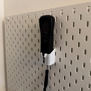 May include: A black video game controller is mounted in a white holder on a white pegboard. The controller has a black wrist strap. The pegboard has evenly spaced oval holes.