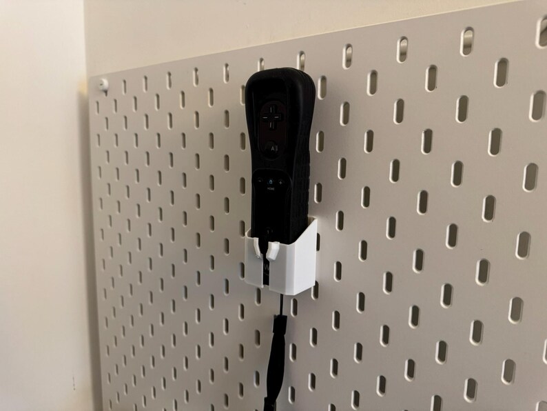 May include: A black video game controller is mounted in a white holder on a white pegboard. The controller has a black wrist strap. The pegboard has evenly spaced oval holes.