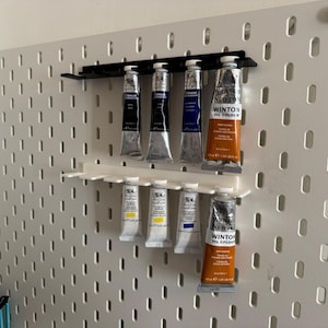 Pode incluir: Um painel perfurado branco com suportes para tubos de tinta pretos e brancos. Os suportes exibem vários tubos de tinta a óleo em várias cores, incluindo azul, amarelo e laranja. Os tubos estão rotulados como "Winton Oil Colour" e "Winsor & Newton."