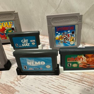 May include: A collection of retro video game cartridges, including titles like "Golf," "Super Mario Land," "Finding Nemo," and "That's So Raven 2." The cartridges are in various colors and designs, with labels featuring game titles and artwork.