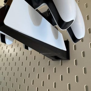 K&ouml;nnte beinhalten: Ein schwarz-wei&szlig;er Gaming-Controller und eine Ladestation, montiert auf einer beigefarbenen Lochwand. Der Controller ist wei&szlig; mit schwarzen Akzenten. Die Ladestation ist schwarz mit wei&szlig;en Oberfl&auml;chen.