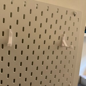 以下が含まれることがあります： 白いペグボードに、白いプラスチック製のフックが2つ取り付けられています。ペグボードには、穴のグリッドパターンがあります。