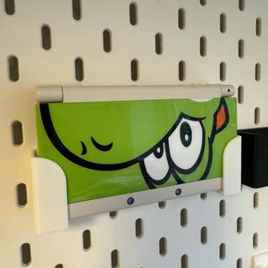 Puede incluir: Una consola de juegos port&aacute;til blanca y verde con un dise&ntilde;o de ojo de dibujos animados est&aacute; montada en una placa perforada blanca. El dispositivo se sujeta con un soporte blanco. La placa perforada tiene agujeros ovalados espaciados.