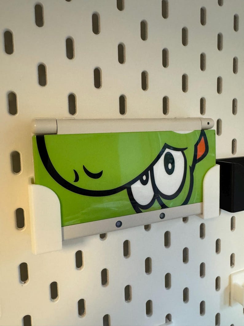 Puede incluir: Una consola de juegos port&aacute;til blanca y verde con un dise&ntilde;o de ojo de dibujos animados est&aacute; montada en una placa perforada blanca. El dispositivo se sujeta con un soporte blanco. La placa perforada tiene agujeros ovalados espaciados.