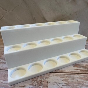 Puede incluir: Un expositor blanco escalonado con tres niveles, cada uno con m&uacute;ltiples hendiduras circulares. El expositor est&aacute; hecho de un material s&oacute;lido y opaco, dise&ntilde;ado para sostener objetos peque&ntilde;os. El fondo es una superficie clara y texturizada.