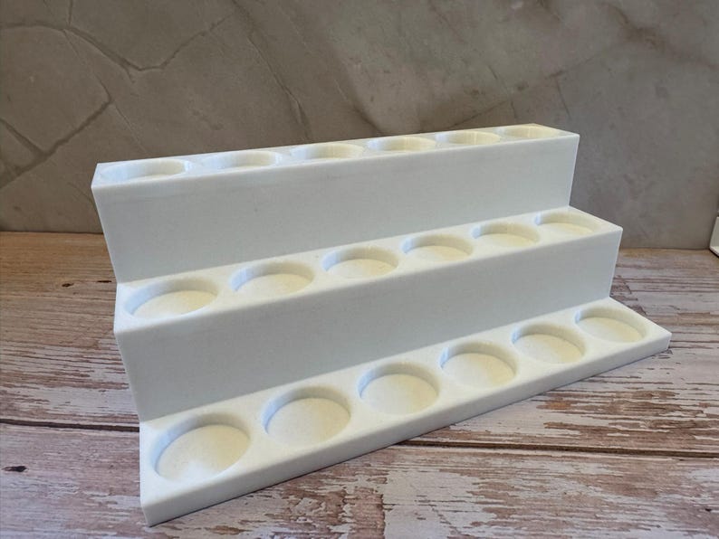 Puede incluir: Un expositor blanco escalonado con tres niveles, cada uno con m&uacute;ltiples hendiduras circulares. El expositor est&aacute; hecho de un material s&oacute;lido y opaco, dise&ntilde;ado para sostener objetos peque&ntilde;os. El fondo es una superficie clara y texturizada.