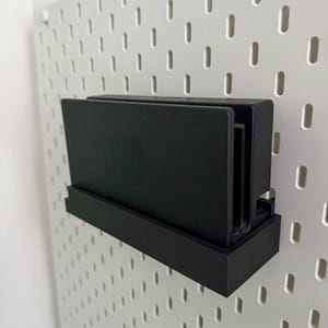 May include: A black Nintendo Switch console and two game cartridges are mounted on a white pegboard wall using a black plastic holder.