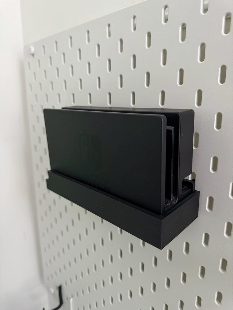 May include: A black Nintendo Switch console and two game cartridges are mounted on a white pegboard wall using a black plastic holder.