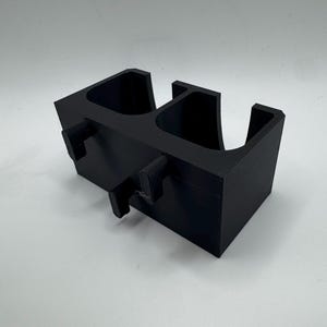 Puede incluir: Un portavasos negro impreso en 3D para coche con dos compartimentos y pesta&ntilde;as de montaje. El soporte es rectangular con bordes redondeados y est&aacute; dise&ntilde;ado para encajar en la consola de un veh&iacute;culo. El portavasos es de pl&aacute;stico.