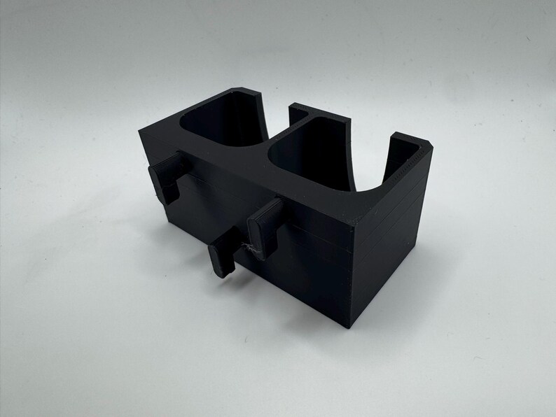 Puede incluir: Un portavasos negro impreso en 3D para coche con dos compartimentos y pesta&ntilde;as de montaje. El soporte es rectangular con bordes redondeados y est&aacute; dise&ntilde;ado para encajar en la consola de un veh&iacute;culo. El portavasos es de pl&aacute;stico.