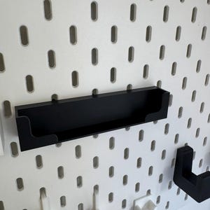 May include: A black shelf attached to a white pegboard. The shelf is rectangular with a raised lip on the front and is designed to hold small items. The pegboard has evenly spaced oval holes.