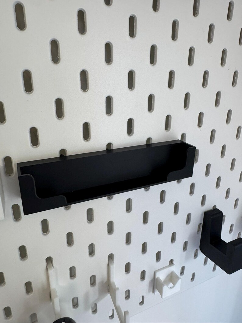 May include: A black shelf attached to a white pegboard. The shelf is rectangular with a raised lip on the front and is designed to hold small items. The pegboard has evenly spaced oval holes.