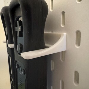 May include: Black game controllers are held by white wall mounts. The controllers have buttons and directional pads. The wall mounts are attached to a wall with rectangular cutouts.
