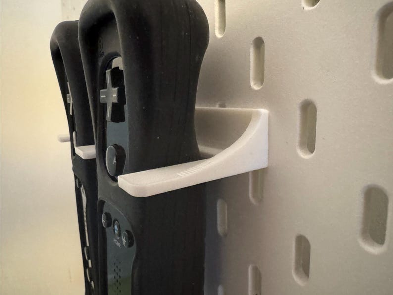 May include: Black game controllers are held by white wall mounts. The controllers have buttons and directional pads. The wall mounts are attached to a wall with rectangular cutouts.