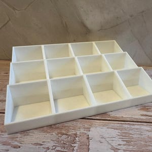 Peut inclure: Un organisateur blanc &agrave; plusieurs niveaux avec de multiples compartiments carr&eacute;s. L'organisateur est fait d'un mat&eacute;riau solide et opaque et est con&ccedil;u pour contenir de petits objets. Il repose sur une surface en bois avec un fond neutre.