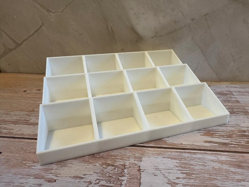 Peut inclure: Un organisateur blanc &agrave; plusieurs niveaux avec de multiples compartiments carr&eacute;s. L'organisateur est fait d'un mat&eacute;riau solide et opaque et est con&ccedil;u pour contenir de petits objets. Il repose sur une surface en bois avec un fond neutre.