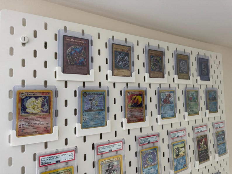 Pu&ograve; includere: Una parete bianca con un pannello forato su cui sono esposte diverse carte da collezione in buste di plastica trasparenti. Le carte sono organizzate in file e colonne, alcune carte presentano personaggi di Pokemon.