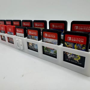 K&ouml;nnte beinhalten: Ein wei&szlig;er, 3D-gedruckter Spielkartenhalter f&uuml;r Nintendo Switch-Spiele. Der Halter hat mehrere Schlitze f&uuml;r Spielkarten und einen Platz f&uuml;r eine Switch-Konsole. Mehrere Spielkarten sind sichtbar, mit dem Nintendo Switch-Logo.