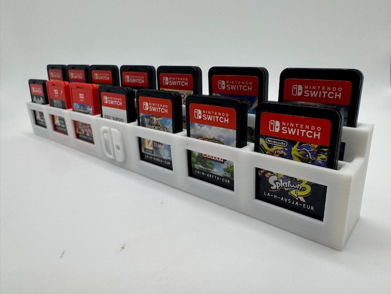 K&ouml;nnte beinhalten: Ein wei&szlig;er, 3D-gedruckter Spielkartenhalter f&uuml;r Nintendo Switch-Spiele. Der Halter hat mehrere Schlitze f&uuml;r Spielkarten und einen Platz f&uuml;r eine Switch-Konsole. Mehrere Spielkarten sind sichtbar, mit dem Nintendo Switch-Logo.