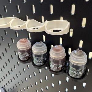 Puede incluir: Cinco peque&ntilde;os frascos de pintura Citadel Colour, en tonos marrones y negros, se muestran en una placa de pared negra perforada. Los frascos se mantienen en su lugar mediante soportes de pl&aacute;stico blanco.