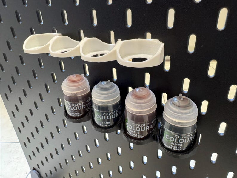 Puede incluir: Cinco peque&ntilde;os frascos de pintura Citadel Colour, en tonos marrones y negros, se muestran en una placa de pared negra perforada. Los frascos se mantienen en su lugar mediante soportes de pl&aacute;stico blanco.