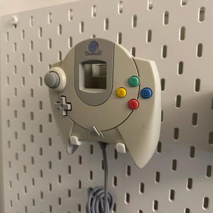 Puede incluir: Un mando Dreamcast beige montado en una pared perforada blanca. El mando tiene una cruceta, una palanca analógica y botones de acción de colores. Un cable gris cuelga por debajo. El logotipo de Dreamcast es visible.