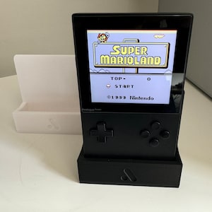 May include: Black handheld gaming console showing "Super Marioland" on screen. The console has a directional pad, buttons, and a stand. A white stand is to the left. The game's title and Nintendo copyright are visible.
