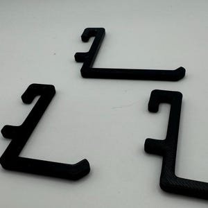 May include: Three black plastic hooks, each shaped like an 'L', are arranged on a white surface.