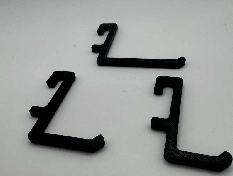 May include: Three black plastic hooks, each shaped like an 'L', are arranged on a white surface.