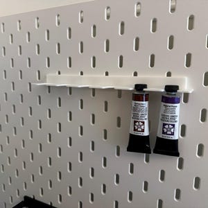 May include: A white pegboard wall organizer featuring a horizontal rack with multiple slots. Two tubes of Daniel Smith Extra Fine Watercolors, specifically Burnt Orange and Imperial Purple, are hanging from the rack by their crimped ends.