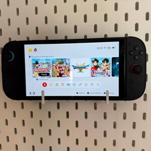 May include: A black Nintendo Switch console displaying a game selection screen, mounted on a white pegboard. The screen shows various game icons, including Monopoly and Mario games. The console is held in place by white brackets.