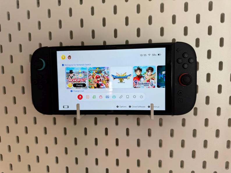 May include: A black Nintendo Switch console displaying a game selection screen, mounted on a white pegboard. The screen shows various game icons, including Monopoly and Mario games. The console is held in place by white brackets.