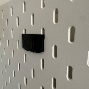 K&ouml;nnte beinhalten: Ein schwarzes Lochwandzubeh&ouml;r ist an einer wei&szlig;en Lochwand befestigt. Die Lochwand hat eine Reihe von gleichm&auml;&szlig;ig verteilten, l&auml;nglichen L&ouml;chern. Das Zubeh&ouml;r scheint ein Halter oder eine Halterung zu sein.