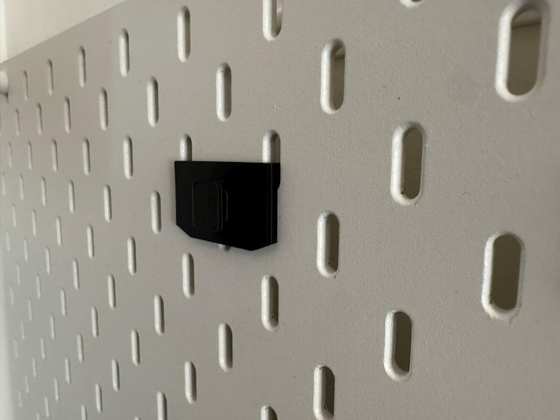 K&ouml;nnte beinhalten: Ein schwarzes Lochwandzubeh&ouml;r ist an einer wei&szlig;en Lochwand befestigt. Die Lochwand hat eine Reihe von gleichm&auml;&szlig;ig verteilten, l&auml;nglichen L&ouml;chern. Das Zubeh&ouml;r scheint ein Halter oder eine Halterung zu sein.