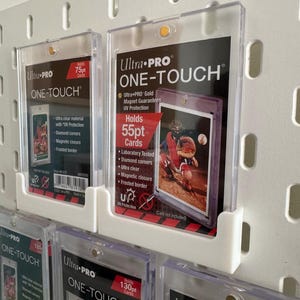 May include: Clear Ultra PRO ONE-TOUCH card holders, designed to protect trading cards. The holders are displayed on a white pegboard. The labels indicate the holders can hold 55pt, 75pt, and 130pt cards. Features include UV protection and magnetic closure.