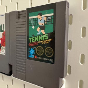 Pode incluir: Dois cartuchos de videojogos retro. O cartucho da direita tem o r&oacute;tulo "Tennis" com um gr&aacute;fico pixelizado de um jogador de t&eacute;nis. O outro cartucho tem o r&oacute;tulo "Goal!" com um gr&aacute;fico de um jogador de futebol. Ambos s&atilde;o cinzentos e montados numa parede perfurada branca.