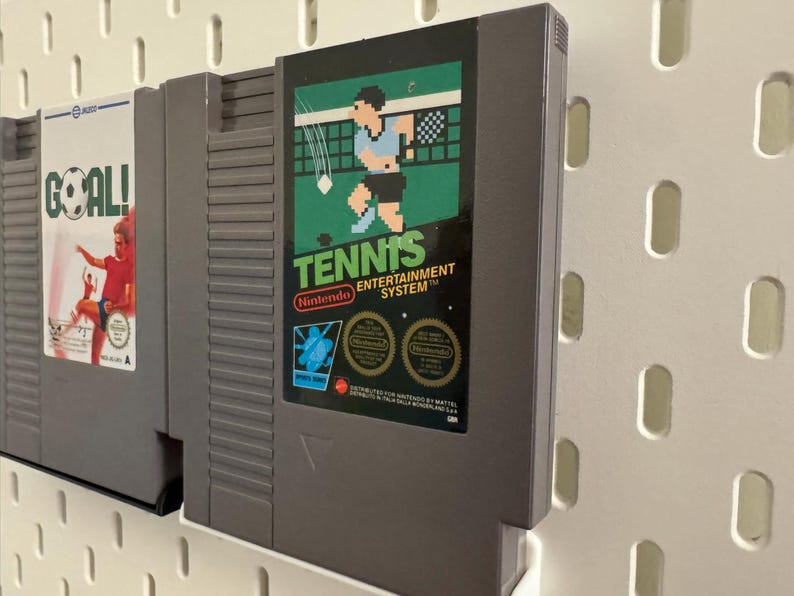Pode incluir: Dois cartuchos de videojogos retro. O cartucho da direita tem o r&oacute;tulo "Tennis" com um gr&aacute;fico pixelizado de um jogador de t&eacute;nis. O outro cartucho tem o r&oacute;tulo "Goal!" com um gr&aacute;fico de um jogador de futebol. Ambos s&atilde;o cinzentos e montados numa parede perfurada branca.