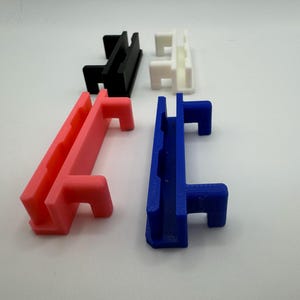 Pode incluir: Quatro suportes de parede de pl&aacute;stico impressos em 3D em diferentes cores: preto, branco, rosa e azul. Cada suporte tem duas ranhuras e um gancho.