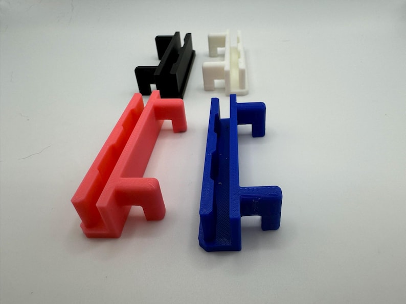 Pode incluir: Quatro suportes de parede de pl&aacute;stico impressos em 3D em diferentes cores: preto, branco, rosa e azul. Cada suporte tem duas ranhuras e um gancho.