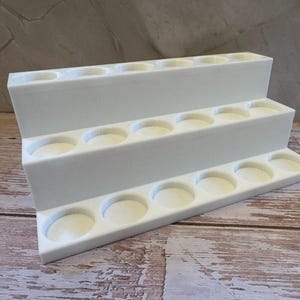 Puede incluir: Un expositor blanco escalonado con tres niveles, cada uno con m&uacute;ltiples huecos circulares. El expositor est&aacute; dise&ntilde;ado para sostener objetos peque&ntilde;os y est&aacute; sobre una superficie de madera.