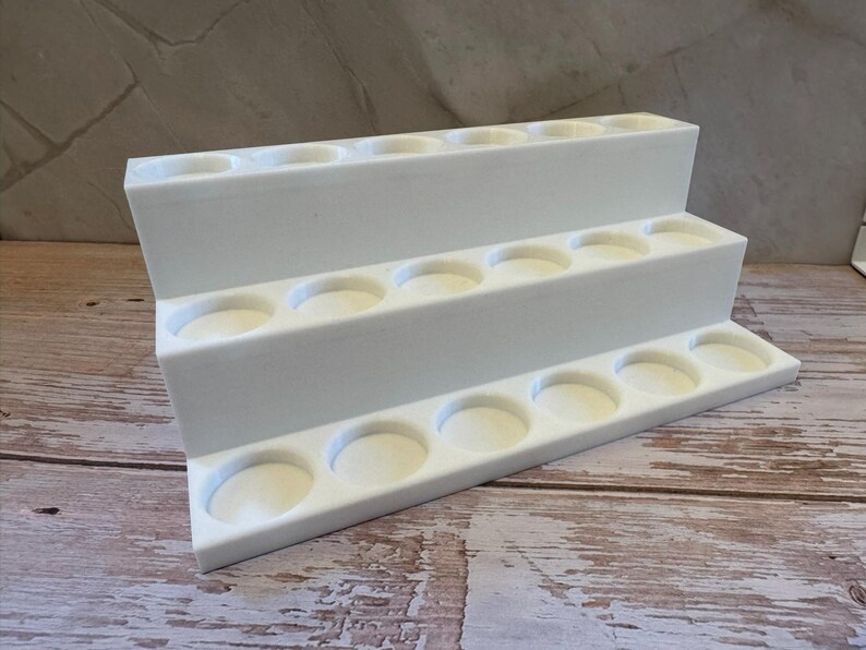 Puede incluir: Un expositor blanco escalonado con tres niveles, cada uno con m&uacute;ltiples huecos circulares. El expositor est&aacute; dise&ntilde;ado para sostener objetos peque&ntilde;os y est&aacute; sobre una superficie de madera.