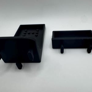 May include: Two black plastic components, likely parts of a larger device, are displayed against a white background. One is a larger, angled piece with several small circular indentations. The other is a smaller, rectangular piece with two protruding pegs.