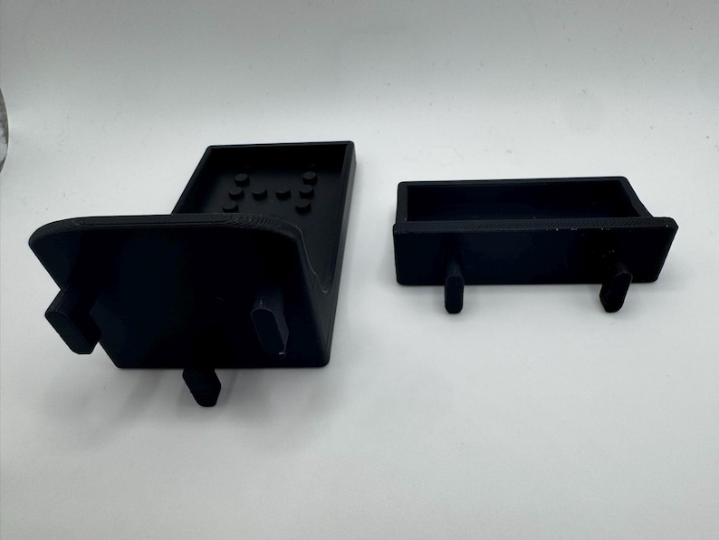 May include: Two black plastic components, likely parts of a larger device, are displayed against a white background. One is a larger, angled piece with several small circular indentations. The other is a smaller, rectangular piece with two protruding pegs.