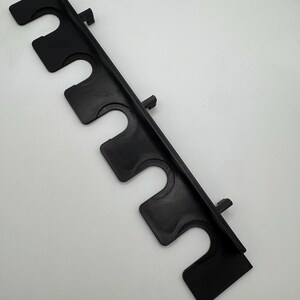 May include: A black plastic rod holder featuring five U-shaped slots. The holder has a textured surface and is designed to hold rods or similar items. The holder is angled and has a rectangular shape.