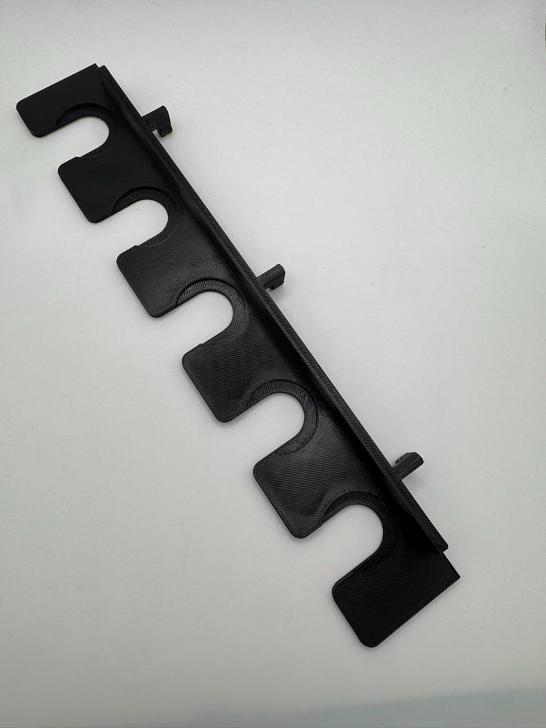 May include: A black plastic rod holder featuring five U-shaped slots. The holder has a textured surface and is designed to hold rods or similar items. The holder is angled and has a rectangular shape.