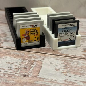 Puede incluir: Un soporte para cartuchos de juego impreso en 3D en blanco y negro con varios cartuchos de juego de Nintendo 3DS y DS. El juego de 3DS presenta Super Mario Maker 3DS. El juego de DS es Dr. Kawashima's Brain Training.