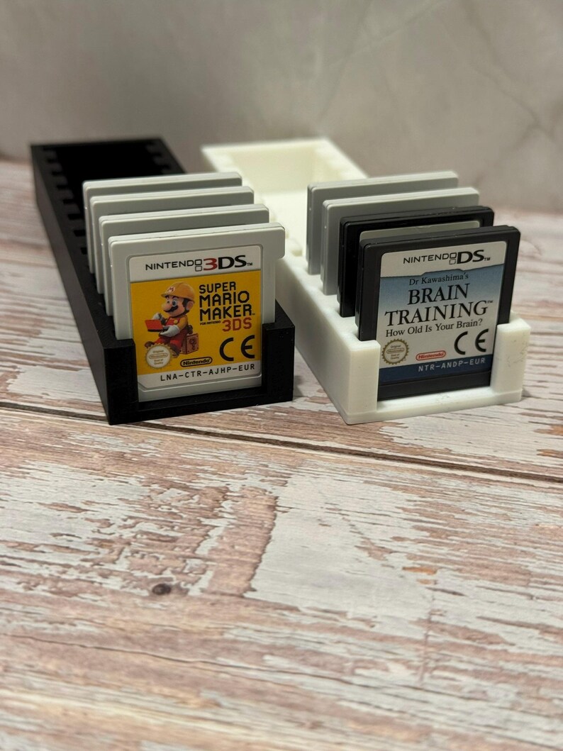 Puede incluir: Un soporte para cartuchos de juego impreso en 3D en blanco y negro con varios cartuchos de juego de Nintendo 3DS y DS. El juego de 3DS presenta Super Mario Maker 3DS. El juego de DS es Dr. Kawashima's Brain Training.